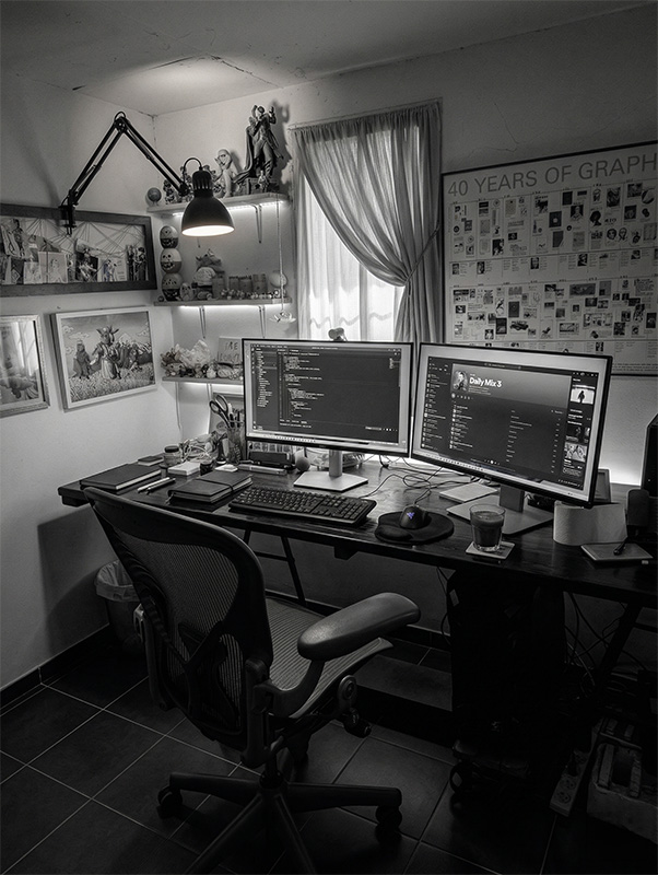 YuvalDesign studio — dual-monitor workstation with design work on screens, architectural lamp, figurines and a 40 Years of Graphic Design poster on the wall